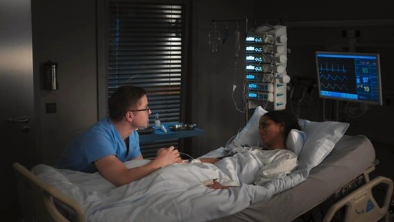 Ein Mann sitzt am Krankenbett einer jungen Frau und hält ihre Hand, sie schauen sich an, die Frau lächelt. Über ihr hängt ein Bildschirm, der medizinische Daten anzeigt. © Screenshot 