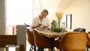 Eine blonde Frau sitzt an einem Tisch in einer hellen Wohnung und füllt Formulare aus. © Screenshot 