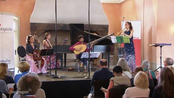 Bei einem Konzert in einem Veranstaltungssaal spielen eine Flötistin und weitere Musiker auf einer kleinen Bühne vor Zuschauern. © Screenshot 