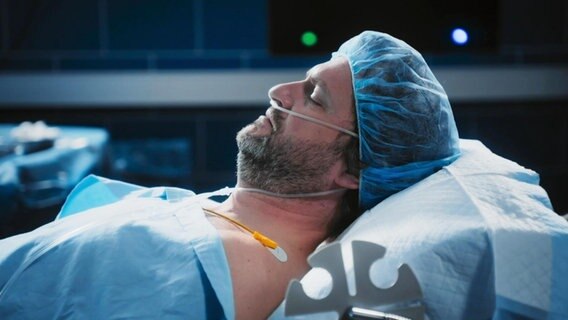 Ein Mann liegt mit geschlossenen Augen auf einem OP-Tisch. Er trägt ein Haarnetz und hat einen Beatmungsschlauch in der Nase. © Screenshot 