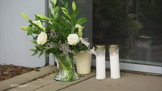 Blumen und Grabkerzen stehen als Erinnerung für zwei verstorbene Mitarbeiter vor einer Firmentür. © Screenshot 