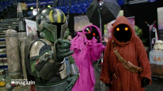 Ein Cosplayer in einer Mandalorian-Rüstung mit zwei Cosplayern in Kapuzenkleidung der Jawas aus Star Wars © Screenshot 