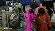 Ein Cosplayer in einer Mandalorian-Rüstung mit zwei Cosplayern in Kapuzenkleidung der Jawas aus Star Wars © Screenshot 