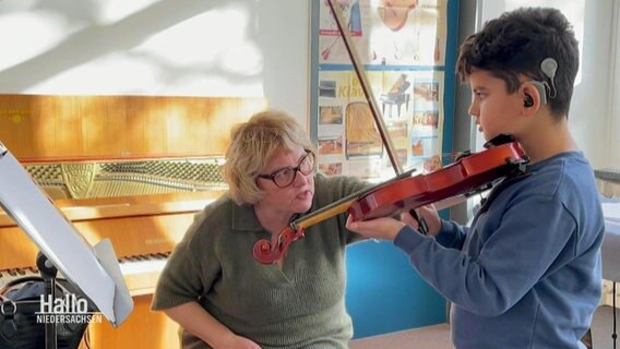 Ein Junge mit Hörgerät spielt Geige und seine Lehrerin sitz neben ihm. © Screenshot 