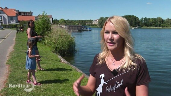 Eine junge Frau im Interview, im Hintergrund stehen eine Mutter mit ihrer Tochter angelnd an einem See. © Screenshot 