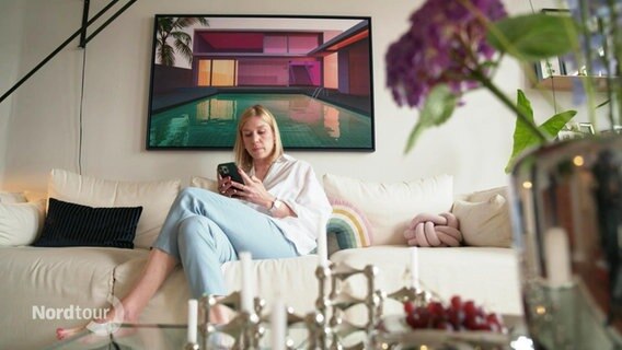 Eine Frau sitzt in einem Wohnzimmer und schaut auf ihr Handy. © Screenshot 