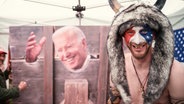Ein Schamane mit einem Büffelfell auf dem Kopf, vor Joe Biden am Pranger. © NDR 