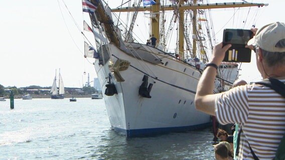 Ein Mann macht mit seinem Handy ein Foto von einem großen Segelschiff im Rostocker Hafen. © Screenshot 