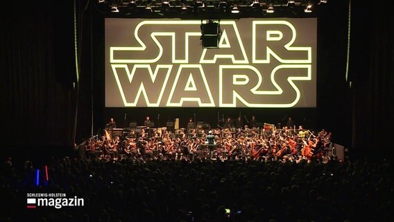 Dunkler Raum mit angeleuchtetem Orchester und auf der Leinwand oberhalb die Aufschrift Star Wars. © Screenshot 