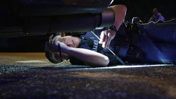 Ein Polizist untersucht mit einer Taschenlampe die Unterseite eines Autos. © Screenshot 