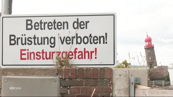 Im Vordergrund ein Schild das "Betreten der Brüstung verboten! Einsturzgefahr" liest, vor dem zu kippen drohenden Leuchtturm in Bremerhaven im Hintergrund. © Screenshot 