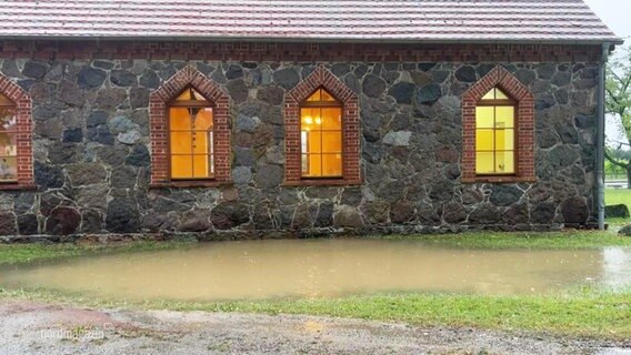 Vor einem älteren Steinhaus befindet sich eine grlßere Pfütze mit bräunlichem Regenwasser. © Screenshot 