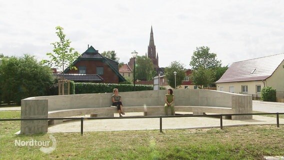 Zwei Frauen sitzen auf einer großen, elliptisch geformten Bank aus Beton. © Screenshot 