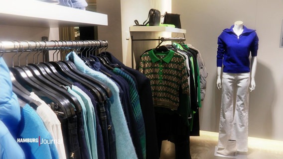 Blick in ein Kleidungsgeschäft in de mehrere Klamotten auf Bügeln aufgehängt sind, vor einer Wand steht eine kopflose Schaufensterpuppe. © Screenshot 