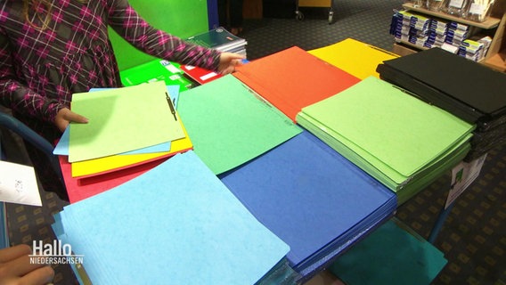 Auf einem Tisch liegen mehrere Stapel verschieden farbiger Schulmappen von denen sich ein kleines Mädchen mehrere auswählt. © Screenshot 