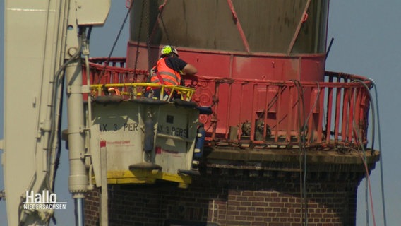 In einer Krankapsel steht ein Arbeiter, sodass er nah an der roten Kuppel des Molenturms in Bremerhaven arbeiten kann. © Screenshot 