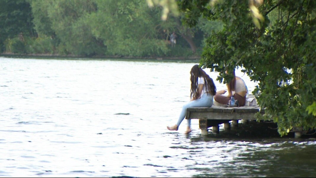 An einem See spielen zwei Personen, die auf einem Steg sitzen, mit den Füßen im Wasser.