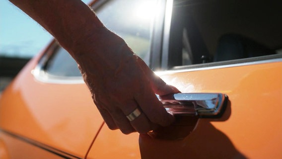 Eine Hand greift an die Türklinke eines orangefarbenen Autos. © Screenshot 