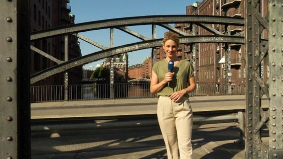 Moderatorin Eva Diederich steht mit Mikrophon auf der Poggenmühlen-Brücke in der Hamburger Speicherstadt. © Screenshot 