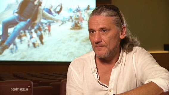 Regisseur Mihail Sofronea im Interview. Hinter ihm eine Leinwand, auf der ein Film läuft. © Screenshot 