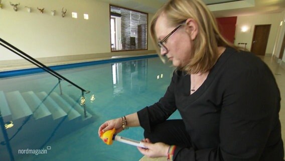 Eine Frau begutachtet ein Thermometer mit dem sie die Temperatur eines Indoor-Schwimmbeckens gemessen hat. © Screenshot 