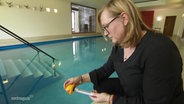 Eine Frau begutachtet ein Thermometer mit dem sie die Temperatur eines Indoor-Schwimmbeckens gemessen hat. © Screenshot 