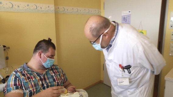 Ein Arzt schaut in einem Krankenhauszimmer zu einem Vater, der seinem Baby zu trinken gibt. © Screenshot 