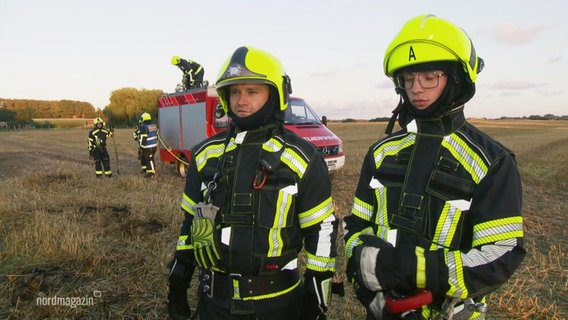 Zwei Feuerwehrmänner stehen auf einem Feld mit ihrer neuen Schutzkleidung, im Hintergrund: ein Einsatzwagen mit weiteren Feuerwehrleuten © Screenshot 