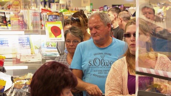 Viele Leute stehen bei einer Supermarktkasse in einer Schlange, einige sehen etwas genervt aus. © Screenshot 