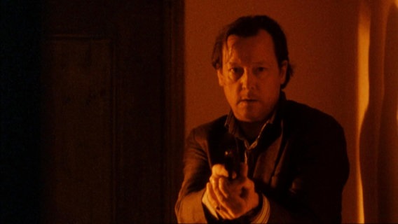 Hauptkommissar Hanns von Meuffels (gespielt von Matthias Brandt) geht mit gezogener Waffe durch einen Flur, die Atmosphäre ist in ein rötliches Licht getaucht. © Screenshot 
