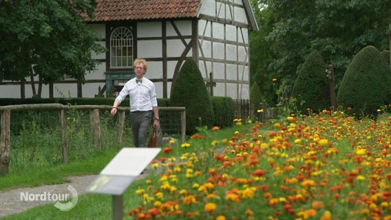 Ein Mann geht einen kleinen Feldweg an einer Blumenwiese entlang, im Hintergrund: ein Fachwerkhaus. © Screenshot 