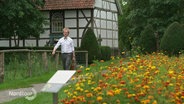 Ein Mann geht einen kleinen Feldweg an einer Blumenwiese entlang, im Hintergrund: ein Fachwerkhaus. © Screenshot 