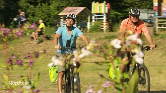 Zwei Fahrradfahrer fahren auf einem sonnigen Feldweg entlang. © Screenshot 