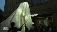 In einer Fußgängerzone wird bei abendlicher Dämmerung eine überdimensionierte, beleuchtete Statue mit Stöckern zur Bewegung gebracht. © Screenshot 