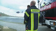 Zwei Feuerwehrleute stehen mit dem Rücken zur Kamera. Sie blicken auf einen See, an dem ein Löschhubschrauber Wasser auftankt. © Screenshot 