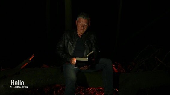 Autor Andreas Winkelmann sitzt bei tiefer Nacht auf einem Baumstamm und liest aus einem seiner Bücher vor. Er hält dabei eine Taschenlampe als Lichtquelle. © Screenshot 