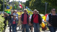 Eine Anzahl von Menschen zieht als Demonstrationszug über eine Straße. Sie halten Banner hoch oder auch die deutsche Flagge. © Screenshot 