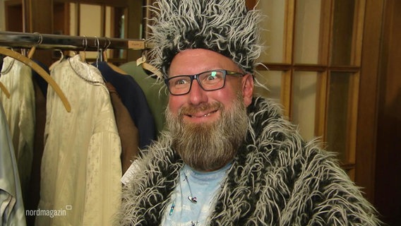 Ein Mann schaut breit grinsend in die Kamera. Er trägt einen Pelzmantel samt passender Mütze, an dem noch längere weiße Haare hängen. © Screenshot 
