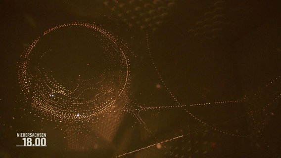 Eine Lichtinstallation wirft goldene Punkte an die Wände eines dunklen Raumes. © Screenshot 
