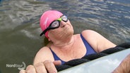 Eine ältere Dame mit rosa Schwimmkappe und Schwimmbrille hält sich im Wasser an einem Bootsrand fest. © Screenshot 