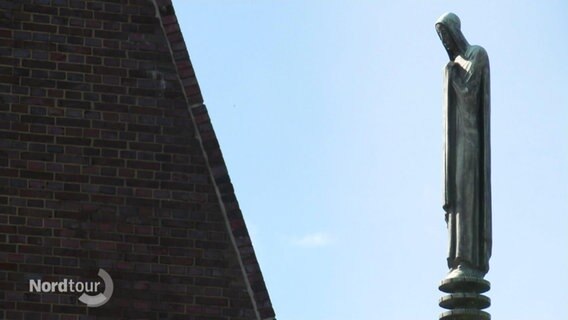 Eine längliche, dürre Statue in Kapuzenkutte steht auf einem Pfahl neben einem Häuserdach. © Screenshot 