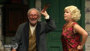 Ausschnitt aus einem Theaterstück: Ein älterer Mann hält sich an ein Höhrrohr ans Ohr, eine blonde Frau stemmt daneben genervt die Hände in die Hüften. © Screenshot 