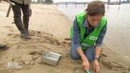 Eine Frau kniet im Sand am Strand mit einer grünen Warnweste und schaufelt Sanproben in eine Metallbrotdose. © Screenshot 