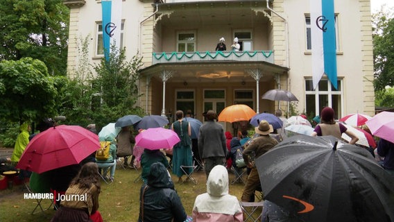 Zuschauer stehen vor einem Balkon, auf dem eine Szene gespielt wird. © Screenshot 