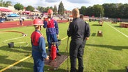 Zwei Kinder in Feuerwehruniform und ein Schiedsrichter stehen bei einem Wettbewerb auf einem Sportplatz. © Screenshot 
