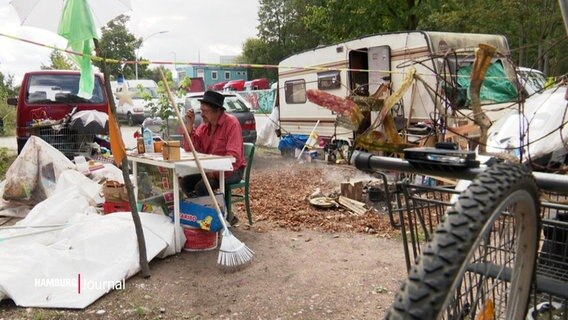 Der Obdachlose Stefan sitzt an einem Tisch, den er auf einer freien Fläche im Freien aufgestellt hat. Um ihn herum stehen diverse Einrichitungsgenstände und im Hintergrund ist ein alter Camper zu sehen. © Screenshot 