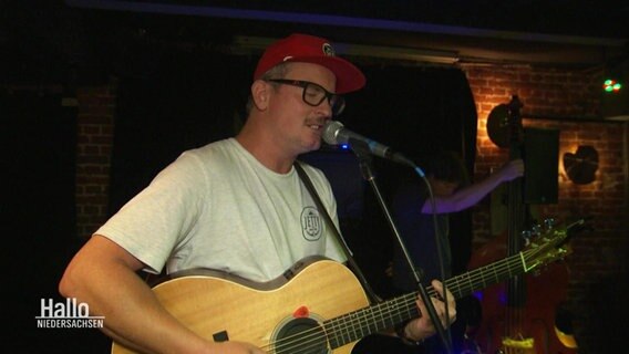 Ein Mann mit Brille und roter Cap steht auf einer Bühne in einem Kulturkeller, spielt Gitarre und singt dazu beherzt in ein Standmikro. © Screenshot 