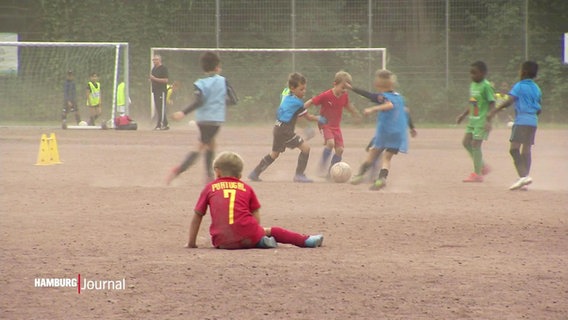 Ein Junge sitzt für eine kurze Pause auf dem Grandplatz eines Sportvereins. Vor ihm spielen weitere Kinder Fußball. Über der Szene steigt sichtbar Staub vom Platz empor. © Screenshot 