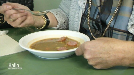 Eine Person sitzt an einem Tisch und löffelt eine Suppe mit Wurststücken darin. © Screenshot 