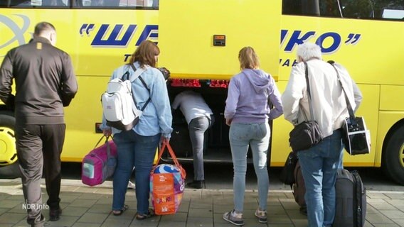 Menschen laden Reisetaschen in einen Bus. © Screenshot 